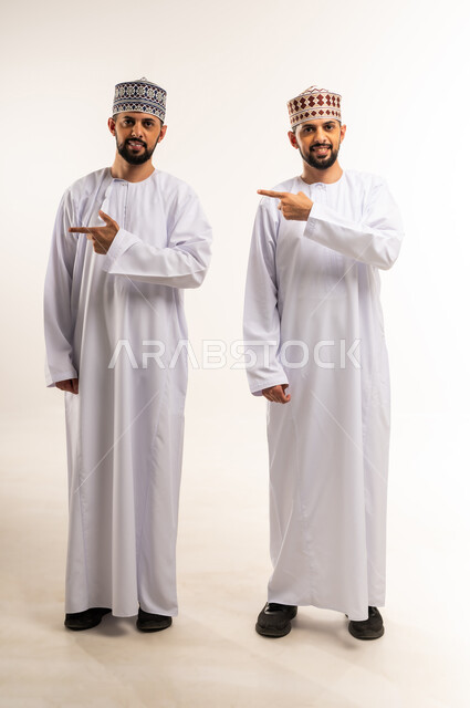 الدلالة على موقع معين, حركات تدل على هذا المكان (هنا), تحديد مكان عنصر محدد, بورتريه لشابين توأم عربيين خليجيين عمانيين يرتديان الدشداشة والكمة يشيران بإصبع السبابة إلى شيء ما, الجسم بالطول الكامل, خلفية بيضاء