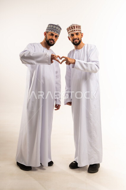 النظر الى الكاميرا بتعابير السرور، تقوية وتعزيز الروابط العائلية، مفهوم الصداقة والأخوة، المحبة والمودة بين الأخوات، بورتريه لشابين توأم عربيين خليجيين عمانيين مبتسمين يرتديان الدشداشة والكمة يرفعان يديهما ويشكلان قلب حب, الجسم بالطول الكامل، خلفية بيضاء