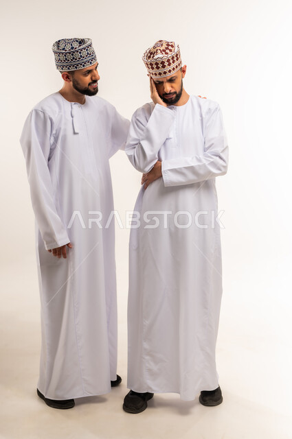 مفهوم التعاطف والمودة بين الأخوات, الوقوف بجانب بعضهم البعض في الأوقات الصعبة, الشعور بالحزن و الاكتئاب, بورتريه لشاب عربي خليجي عماني يرتدي الدشداشة والكمة يضع يده على كتف أخيه التوأم لتخفيف الألم والمواساة, الجسم بالطول الكامل، خلفية بيضاء