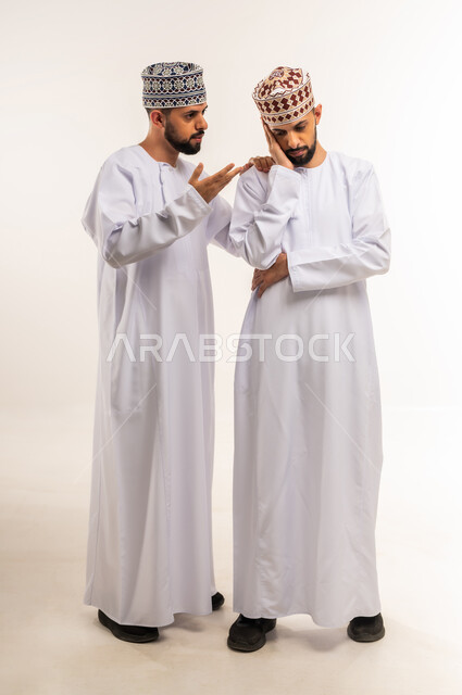 الشعور بالحزن و الاكتئاب، مفهوم التعاطف والمودة بين الأخوات, الوقوف بجانب بعضهم البعض في الأوقات الصعبة, بورتريه لشاب عربي خليجي عماني يرتدي الدشداشة والكمة يضع يده على كتف أخيه التوأم لتخفيف الألم والمواساة, الجسم بالطول الكامل، خلفية بيضاء