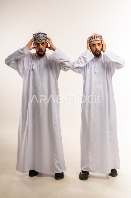 المفاجأة بسماع خبر ما, الشعور بالدهشة والذهول, بورتريه لشابين توأم عربيين خليجيين عمانيين يرتديان الدشداشة والكمة يضعان يديهما على رأسهم بتعابير الصدمة, الجسم بالطول الكامل, خلفية بيضاء