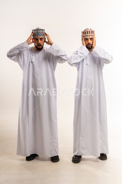 الشعور بالدهشة والذهول, المفاجأة بسماع خبر ما, بورتريه لشابين توأم عربيين خليجيين عمانيين يرتديان الدشداشة والكمة يضعان يديهما على رأسهم بتعابير الصدمة, الجسم بالطول الكامل, خلفية بيضاء