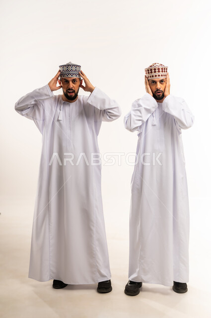 المفاجأة بسماع خبر ما, الشعور بالدهشة والذهول, بورتريه لشابين توأم عربيين خليجيين عمانيين يرتديان الدشداشة والكمة يضعان يديهما على رأسهم بتعابير الصدمة, الجسم بالطول الكامل, خلفية بيضاء