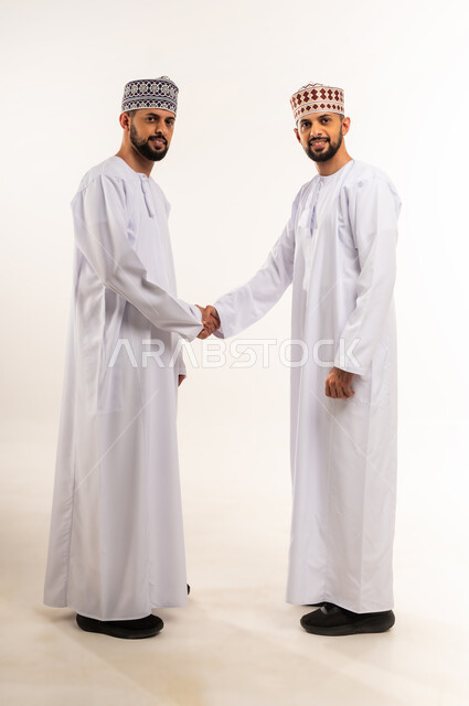 النظر إلى الكاميرا بإيماءات السعادة والسرور, مفهوم المصافحة والاتفاق, بورتريه من الجانب لشابين توأم عربيين خليجيين عمانيين يرتديان الدشداشة والكمة يصافحان بعضهما, الجسم بالطول الكامل, خلفية بيضاء
