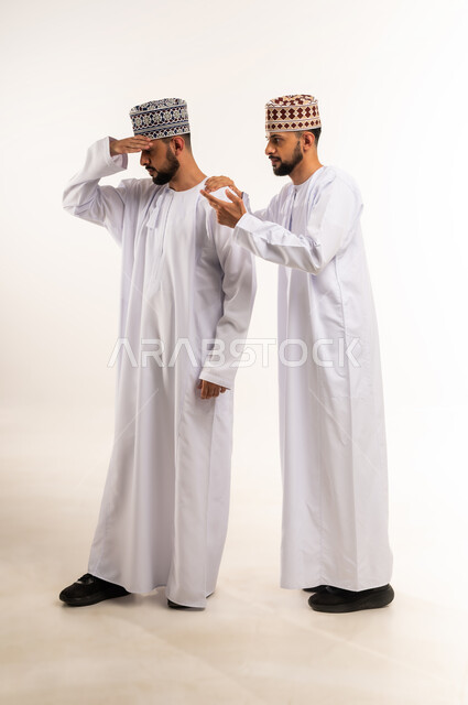 مفهوم التعاطف والمودة بين الأخوات, الوقوف بجانب بعضهم البعض في الأوقات الصعبة, الشعور بالحزن و الاكتئاب, بورتريه لشاب عربي خليجي عماني يرتدي الدشداشة والكمة يضع يده على كتف أخيه التوأم لتخفيف الألم والمواساة, الجسم بالطول الكامل، خلفية بيضاء