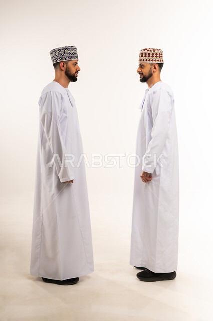 Standing up straight and upright, taking care of one's appearance, the concept of elegance and masculinity, side portrait of two young Omani Gulf Arab twins wearing dishdasha and kummah looking at each other with gestures of happiness and pleasure, full body, white background
