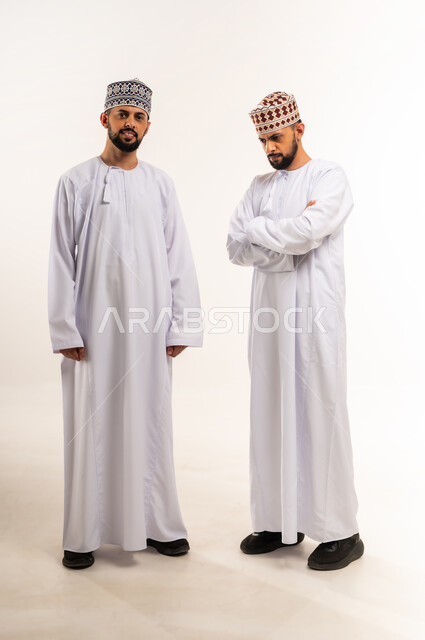 Concern for appearance, concept of elegance and masculinity, looking at the camera with gestures of happiness and pleasure, portrait of two young Omani Arab Gulf twins wearing a dishdasha and a kummah, one standing straight and the other with crossed hands, full body, white background