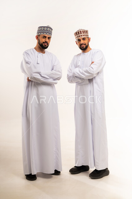 Concept of elegance and masculinity, concern for outward appearance, looking at the camera with gestures of happiness and pleasure, portrait of two young Omani Gulf Arab twins wearing dishdasha and kummah standing straight with crossed hands, full body, white background