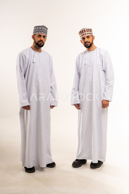 الاهتمام بالمظهر الخارجي, مفهوم الأناقة والرجولة, النظر إلى الكاميرا بإيماءات السعادة والسرور, بورتريه لشابين توأم عربين خليجيين عمانيين يرتديان الدشداشة والكمة يقفان باستقامة واعتدال, الجسم بالطول الكامل, خلفية بيضاء