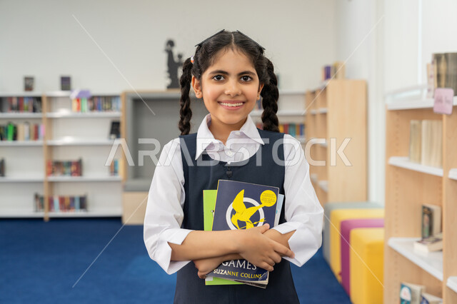 زيارة مكتبة المدرسة، حب الثقافة والاستطلاع، صورة مقربة لطالبة عربية خليجية إماراتية ترتدي الزي المدرسي تحمل مجموعة كتب تنظر إلى الكاميرا بإيماءات السرور والسعادة، التعليم والدراسة في الإمارات، قضاء وقت الفراغ بالتعلم والتثقف الذاتي, تنمية مهارة القراءة للطلاب
