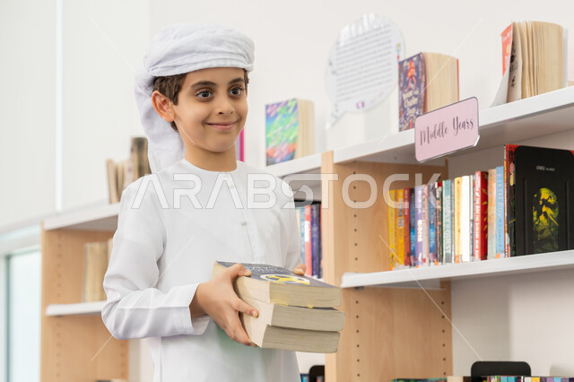 التعليم والدراسة في الإمارات، زيارة مكتبة المدرسة، حب الثقافة والاستطلاع، طالب عربي خليجي إماراتي يرتدي الكندورة والعمامة يحمل بيديه مجموعة من الكتب ينظر إلى شيء ما بإيماءات السرور والسعادة، قضاء وقت الفراغ بالتعلم والتثقف الذاتي, تنمية مهارة القراءة للطلاب