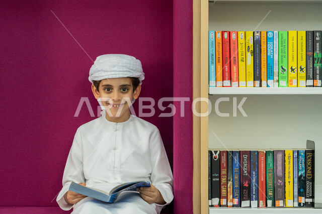 زيارة مكتبة المدرسة، حب الثقافة والاستطلاع، طالب عربي خليجي إماراتي يرتدي الكندورة والعمامة يحمل بيده كتاب ينظر إلى الكاميرا بإيماءات السرور والسعادة، تنمية مهارة القراءة للطلاب، التعليم والدراسة في الإمارات، قضاء وقت الفراغ بالتعلم والتثقف الذاتي