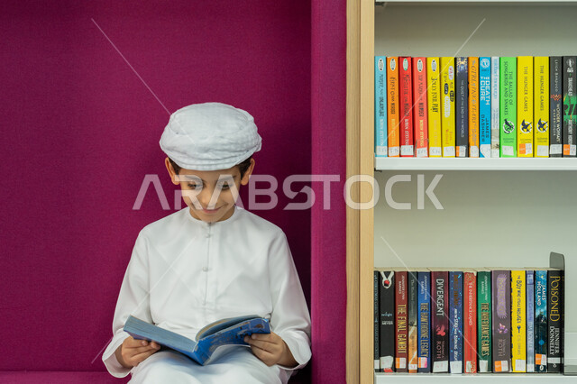 حب الثقافة والاستطلاع، طالب عربي خليجي إماراتي يرتدي الكندورة والعمامة يقوم بقراءة كتاب بإيماءات التركيز والانتباه، تنمية مهارة المطالعة للطلاب، التعليم والدراسة في الإمارات العربية المتحدة، زيارة مكتبة المدرسة، قضاء وقت الفراغ بالتعلم والتثقف الذاتي