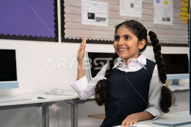 المنهاج المتطور والتعليم الالكتروني في المدرسة، طالبة عربية خليجية إماراتية ترتدي الزي المدرسي ترفع يدها للإجابة عن السؤال، إيماءات الحماس لمعرفة الجواب الصحيح، المشاركة الفعالة للتلاميذ، استخدام جهاز تقني حديث في التعلم، التفاعل والنشاط داخل الفصل الدراسي