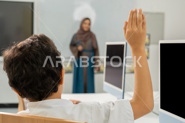 Active student participation, e-learning in school, student raising hand to answer question, Emirati Gulf Arab teacher wearing hijab and abaya standing in front of white board, classroom activity, use of modern technical device in learning, development of teaching method in UAE