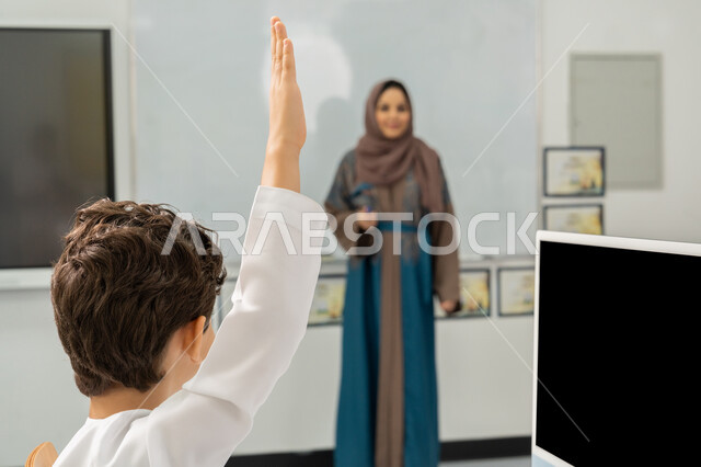 التعليم الالكتروني في المدرسة، طالب يرفع يده للإجابة عن السؤال، معلمة عربية خليجية إماراتية ترتدي الحجاب والعباءة تقف أمام سبورة مدرسية بيضاء، النشاط داخل الفصل الدراسي، استخدام جهاز تقني حديث في التعلم، تطور طريقة التدريس في الإمارات، المشاركة الفعالة للتلاميذ