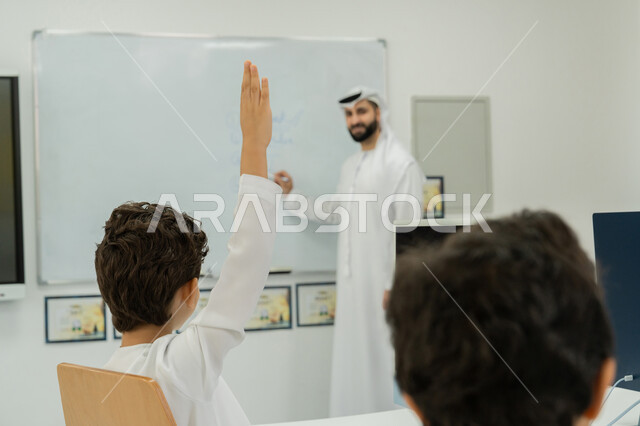 استخدام جهاز تقني حديث في التعلم، التفاعل داخل الفصل الدراسي، المنهاج المتطور والتعليم الالكتروني في المدرسة، معلم عربي خليجي إماراتي يرتدي الكندورة والغترة يشرح الدرس على سبورة مدرسية بيضاء، طالب يرفع يده للإجابة عن السؤال، تطور طريقة التدريس في مدارس الإمارات