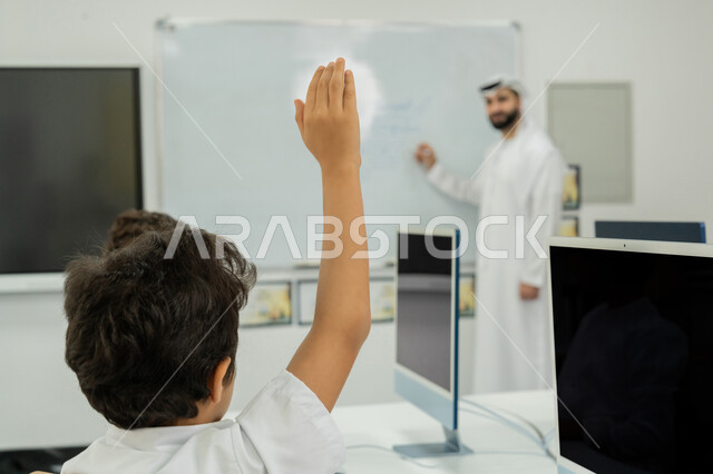 التفاعل داخل الفصل الدراسي، المنهاج المتطور والتعليم الالكتروني في المدرسة، معلم عربي خليجي إماراتي يرتدي الكندورة والغترة يشرح الدرس على سبورة مدرسية بيضاء، طالب يرفع يده للإجابة عن السؤال، استخدام جهاز تقني حديث في التعلم، تطور طريقة التدريس في مدارس الإمارات