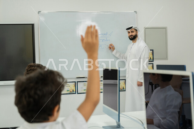 المنهاج المتطور والتعليم الالكتروني في المدرسة، معلم عربي خليجي إماراتي يرتدي الكندورة والغترة يشرح الدرس على سبورة مدرسية بيضاء، طالب يرفع يده للإجابة عن السؤال، التفاعل داخل الفصل الدراسي، استخدام جهاز تقني حديث في التعلم، تطور طريقة التدريس في مدارس الإمارات