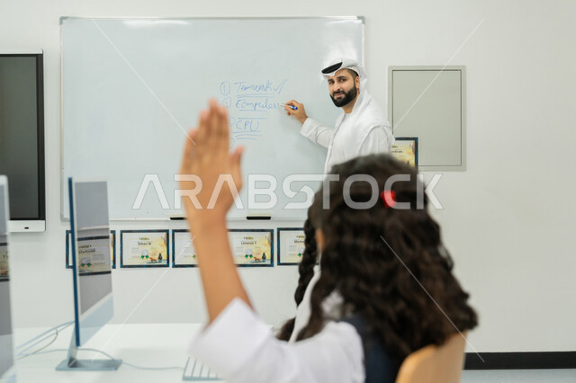 التفاعل والنشاط داخل الفصل الدراسي، التعليم الالكتروني في المدرسة، استخدام جهاز تقني حديث في التعلم، تطور طريقة التدريس في مدارس الإمارات، طالبة إماراتية ترفع يدها للإجابة على السؤال، معلم عربي خليجي إماراتي يرتدي الكندورة والغترة يشرح الدرس على سبورة مدرسية بيضاء