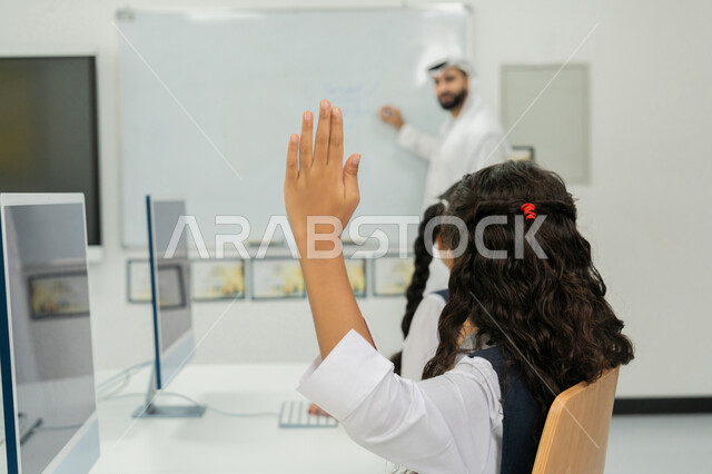 التعليم الالكتروني في المدرسة، استخدام جهاز تقني حديث في التعلم، تطور طريقة التدريس في مدارس الإمارات، طالبة إماراتية ترفع يدها للإجابة على السؤال، معلم عربي خليجي إماراتي يرتدي الكندورة والغترة يشرح الدرس على سبورة مدرسية بيضاء، التفاعل والنشاط داخل الفصل الدراسي