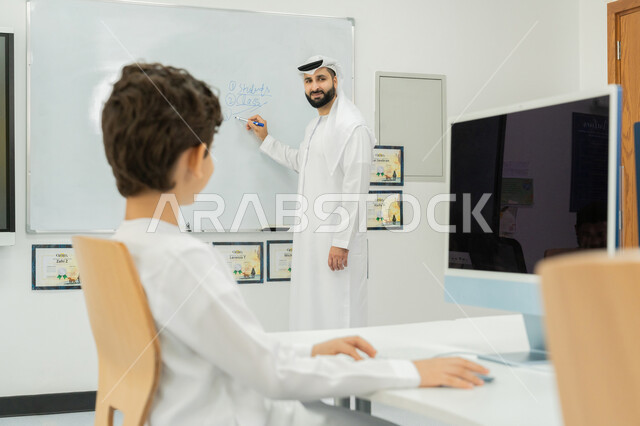 المنهاج المتطور والتعليم الالكتروني في المدرسة، معلم عربي خليجي إماراتي يرتدي الكندورة والغترة يقوم بشرح الدرس على لوحة مدرسية بيضاء، استخدام جهاز تقني حديث في التعلم والدراسة داخل الفصل الدراسي، تطور طريقة التدريس في مدارس الإمارات العربية المتحدة