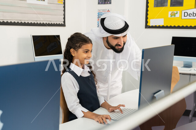 استخدام جهاز تقني حديث في التعلم والدراسة داخل الفصل الدراسي، المنهاج المتطور والتعليم الالكتروني في المدرسة، تطور طريقة التدريس في مدارس الإمارات العربية المتحدة، معلم عربي خليجي إماراتي يرتدي الكندورة والغترة يقوم بشرح الدرس للطالبة باستخدام الحاسوب