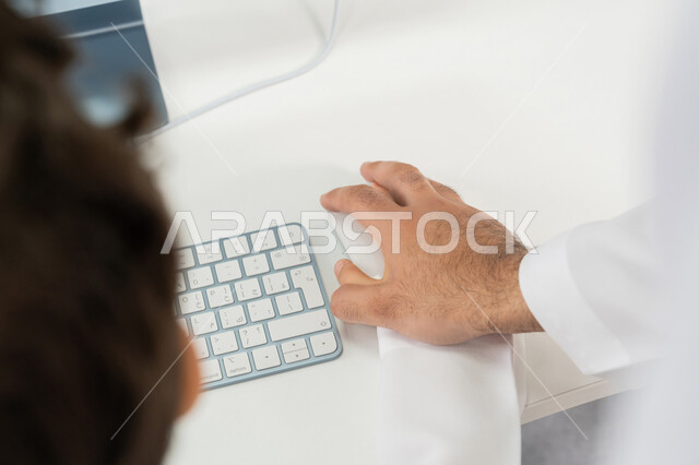 Saudi teacher explaining lesson to student using computer, advanced curriculum and e-learning in school, advanced schools in the Gulf, using modern technical device in learning and studying inside the classroom, close-up of hand of Arab Gulf Emirati teacher wearing kandura using mouse
