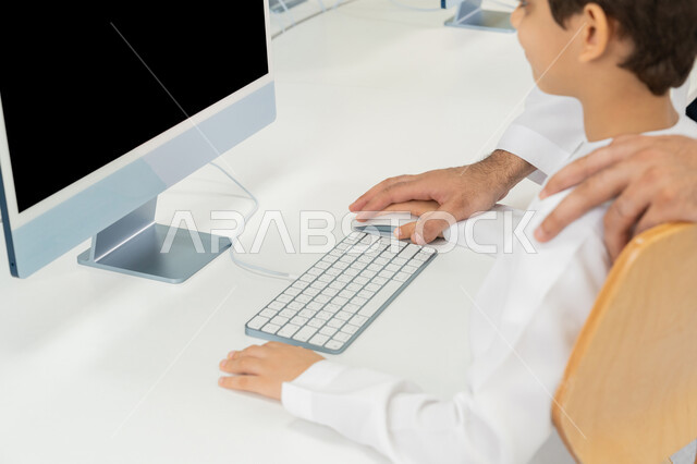 Typing on a keyboard, the development of teaching methods in UAE schools, a close-up of an Emirati Gulf Arab teacher wearing a kandura explaining the lesson to a student using a computer, the advanced curriculum and e-learning in school, the use of a modern technical device in learning and studying inside the classroom
