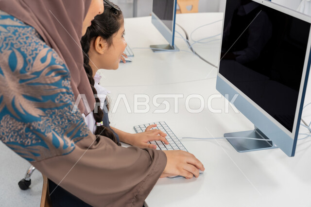 Advanced curriculum and e-learning in school, use of modern technical device in learning and studying inside the classroom, typing on the keyboard, development of teaching method in UAE schools, an Arab Gulf Emirati teacher wearing an abaya explains the lesson to the student using the computer