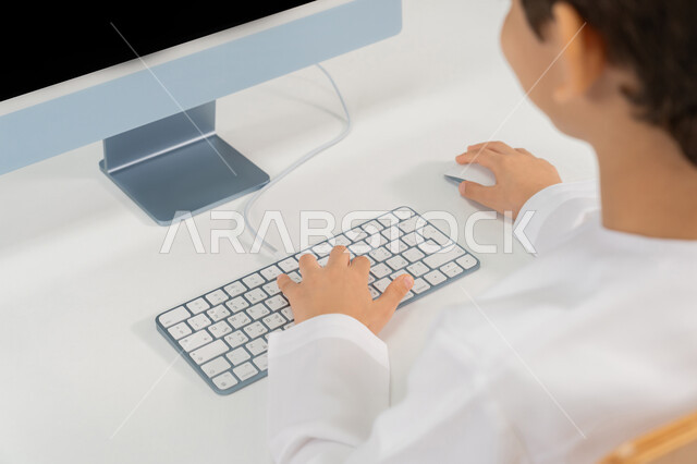 المنهاج المتطور والتعليم الالكتروني في المدرسة، استخدام جهاز تقني حديث  في التعلم والدراسة داخل الفصل الدراسي، الكتابة على لوحة المفاتيح، تطور طريقة التدريس في مدارس الامارات، صورة مقربة من الخلف لطالب عربي خليجي اماراتي يرتدي الكندورة يدرس باستخدام الحاسوب