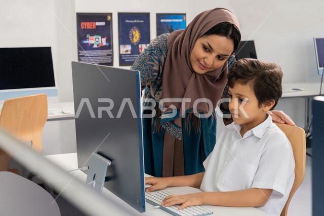 استخدام جهاز تقني حديث في التعلم والدراسة داخل الفصل الدراسي، تطور طريقة التدريس في مدارس الامارات العربية المتحدة، معلمة عربية خليجية اماراتية محجبة ترتدي العباءة تقوم بشرح الدرس لطالب باستخدام الحاسوب المحمول، المنهاج المتطور والتعليم الالكتروني في المدرسة