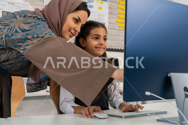 تطور طريقة التدريس في مدارس الامارات العربية المتحدة، معلمة عربية خليجية اماراتية محجبة ترتدي العباءة تقوم بشرح الدرس للطالبة باستخدام الحاسوب، المنهاج المتطور والتعليم الالكتروني في المدرسة، استخدام جهاز تقني حديث  في التعلم والدراسة داخل الفصل الدراسي