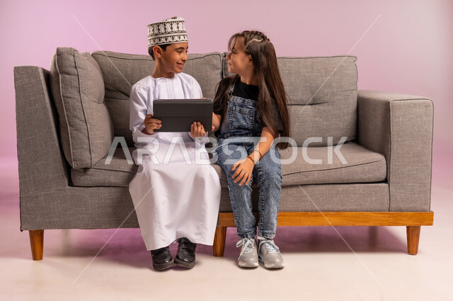 تصفح مواقع التواصل الاجتماعي، التعلم عن بعد، استعمال التكنولوجيا والتقنية الحديثة، بورتريه لولد عربي خليجي عماني يرتدي الدشداشة والكمة يجلس على كنبة مريحة يقوم باستخدام الجهاز اللوحي برفقة أخته، قضاء وقت ممتع بالتسلية على الإنترنت، خلفية وردية