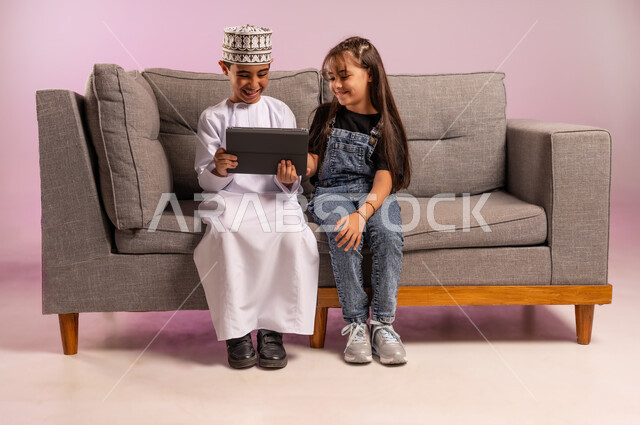 التعلم عن بعد، استعمال التكنولوجيا والتقنية الحديثة، بورتريه لولد عربي خليجي عماني يرتدي الدشداشة والكمة يجلس على كنبة مريحة يقوم باستخدام الجهاز اللوحي برفقة أخته، تصفح مواقع التواصل الاجتماعي، قضاء وقت ممتع بالتسلية على الإنترنت، خلفية وردية