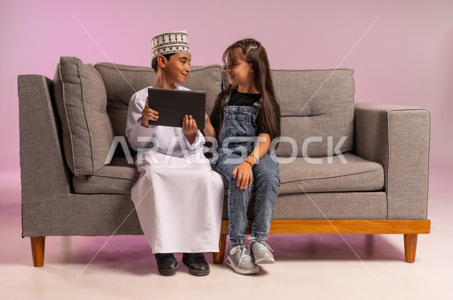 استعمال التكنولوجيا والتقنية الحديثة، بورتريه لولد عربي خليجي عماني يرتدي الدشداشة والكمة يجلس على كنبة مريحة يقوم باستخدام الجهاز اللوحي برفقة أخته، تصفح مواقع التواصل الاجتماعي، قضاء وقت ممتع بالتسلية على الإنترنت، التعلم عن بعد، خلفية وردية