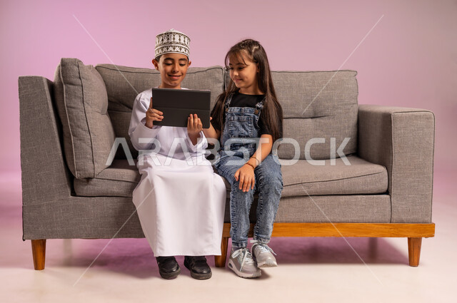 قضاء وقت ممتع بالتسلية على الإنترنت، استعمال التكنولوجيا والتقنية الحديثة، بورتريه لولد عربي خليجي عماني يرتدي الدشداشة والكمة يجلس على كنبة مريحة يقوم باستخدام الجهاز اللوحي برفقة أخته، تصفح مواقع التواصل الاجتماعي، التعلم عن بعد، خلفية وردية