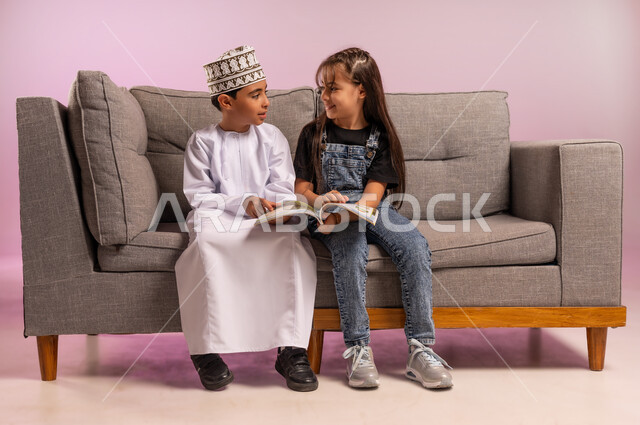 تنمية مهارة القراءة, الاهتمام بتصفح الكتب المفيدة, بورتريه لولد عربي خليجي عماني يرتدي الدشداشة والكمة يجلس على كنبه مريحة يمسك بيده كتاب برفقة أخته, قضاء وقت الفراغ بالتثقيف والمطالعة مع الأصدقاء, مفهوم الثقافة والتعلم الذاتي، خلفية وردية
