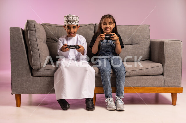 الاستمتاع والتسلية بألعاب الفيديو، بورتريه لولد عربي خليجي عماني يرتدي الدشداشة والكمة يجلس على كنبة مريحة يحمل عصا التحكم بيده يقوم باللعب برفقة اخته بتعابير الحماس والسرور، استخدام التقنية الحديثة في المتعة والترفيه، قضاء وقت ممتع مع الأصدقاء، خلفية وردية