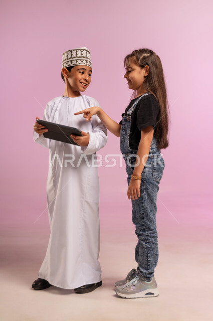 استخدام التكنولوجيا والتقنية الحديثة، بورتريه لولد عربي خليجي عماني يرتدي الدشداشة والكمة يستخدم الجهاز اللوحي برفقة أخته، تصفح مواقع التواصل الاجتماعي، فتاة ترتدي الزي الكاجوال تشير بإصبع السبابة إلى التابلت، الدردشة مع الأصدقاء، الجسم بالطول الكامل، خلفية وردية