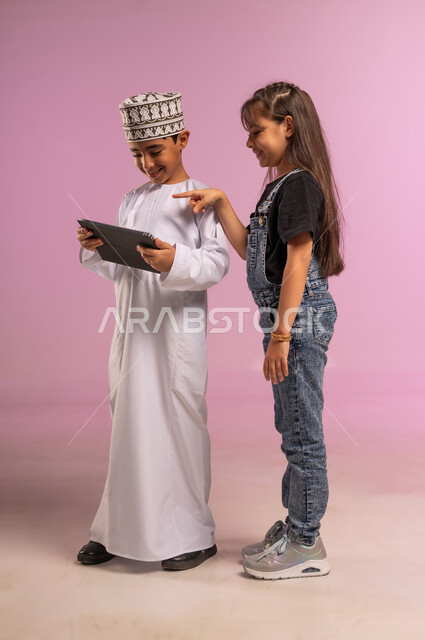 Chatting with friends, using technology and modern technology, portrait of an Arab Gulf Omani boy wearing a dishdasha and a turban using a tablet with his sister, browsing social media, girl in casual wear pointing her index finger at the tablet, full body, pink background