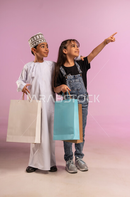 الاستفادة من التخفيض والعرض الموسمي، المشاركة في  التسوق والتبضع مع الاصدقاء، بورتريه لولد عربي خليجي عماني يرتدي الكمة والدشداشة يحمل أكياس تسوق ملونه برفقة اخته، فتاة ترتدي الزي الكاجول تشير بإصبع السبابة إلى شيء ما في الاعلى، الجسم بالطول الكامل، خلفية وردية