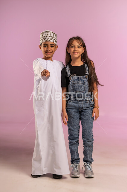 النظر إلى الكاميرا بإيماءات السرور والسعادة، بورتريه لولد عربي خليجي عماني يرتدي الدشداشة والكمة يقف برفقة أخته يشير بإصبع السبابة إلى الأمام لتحديد موقع شيء ما, الجسم بالطول الكامل، خلفية وردية