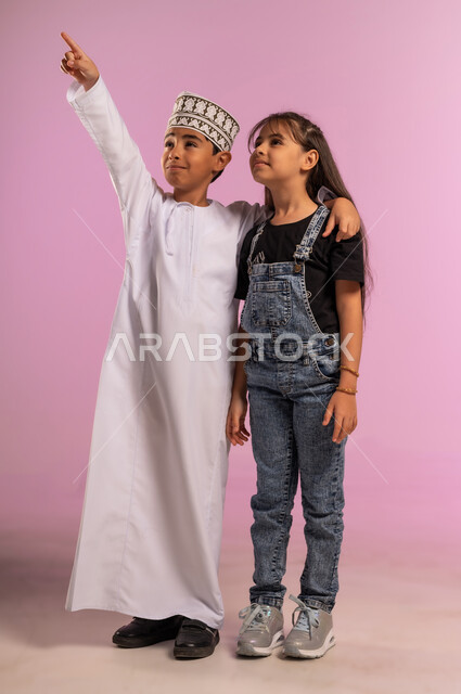 تحديد موقع ومكان عنصر معين، قضاء وقت ممتع مع الأصدقاء، بورتريه لولد عربي خليجي عماني يرتدي الدشداشة والكمة يضع يده على كتف أخته يشير بإصبع السبابة إلى شيء ما في الأعلى, الجسم بالطول الكامل، خلفية وردية