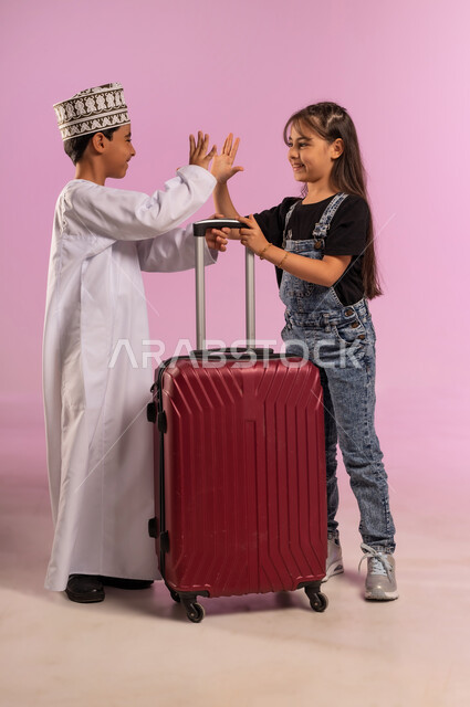 السياحة في إجازة الصيف, الاستعداد لبدء العطلة خارج البلد ,قضاء رحلة صيفية ممتعة، بورتريه لولد عربي خليجي عماني يرتدي الدشداشة والكمة يقف برفقة أخته يمسكان حقيبة السفر يرفعان يدهما بإيماءات السرور والسعادة, الجسم بالطول الكامل، خلفية وردية
