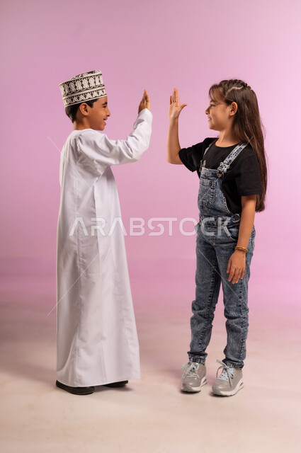 إيماءات ضرب الكف، بورتريه لولد عربي خليجي عماني يرتدي الدشداشة والكمة يرفع يده للأعلى برفقة أخته يلعبان بتعابير السرور والسعادة, قضاء وقت ممتع باللعب مع الأصدقاء، مفهوم الحب والألفة بين الإخوة، الجسم بالطول الكامل، خلفية وردية