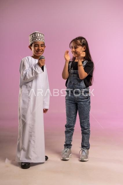 Discovering singing talent since childhood, presenting an artistic show and entertainment segment, portrait of an Arab Gulf Omani boy wearing a dishdasha and a kummah holding a loudspeaker with his sister, recording a singing audio clip, modern technical device, a girl raising her hands clapping with expressions of encouragement, full-length body, pink background