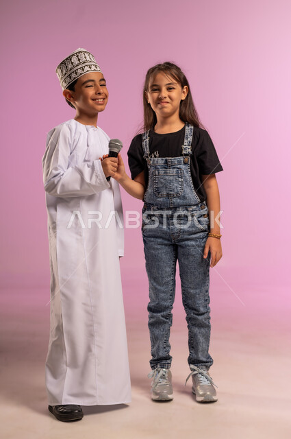 Using a modern technical device, a girl looking at the camera with an expression of pleasure, presenting an artistic show and an entertainment segment, a portrait of an Arab Gulf Omani boy wearing a dishdasha and a kummah holding a loudspeaker with his sister, recording a singing audio clip, discovering the talent for singing since childhood, full-length body, pink background
