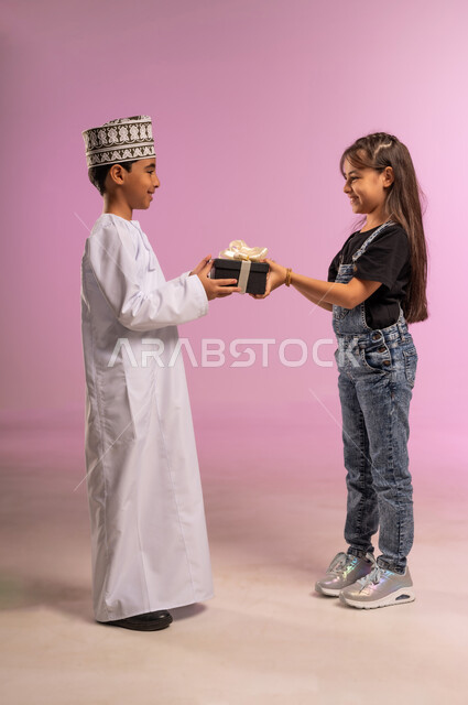 التعبير عن المحبة والتقدير بين الإخوة، مفهوم المفاجأة الخاصة والمناسبة السعيدة, فتاة ترتدي الزي الكاجوال تنظر بتعابير السعادة والسرور، بورتريه لولد عربي خليجي عماني يرتدي الدشداشة والكمة يقوم بتقديم صندوق هدية مغلف إلى أخته، الجسم بالطول الكامل، خلفية وردية