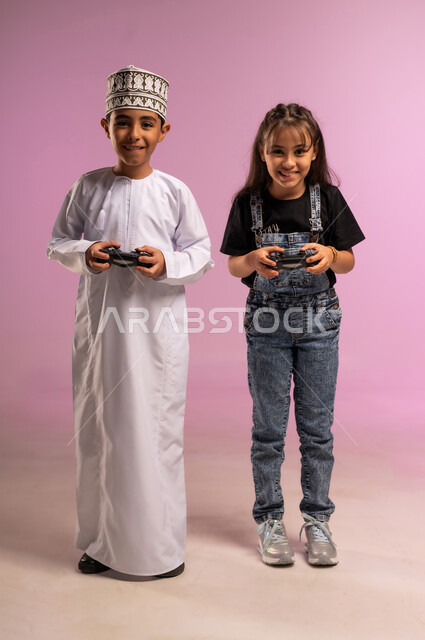Having a good time with friends, enjoying and having fun with video games, portrait of an Arab Gulf Omani boy wearing a dishdasha and a turban, holding a joystick in his hand, playing with his sister, using modern technology for fun and entertainment, expressions of enthusiasm and pleasure, full-length body, pink background